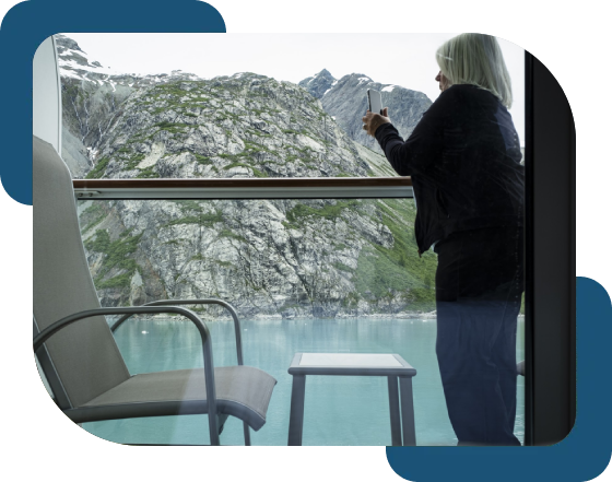 A person stands on a balcony, taking a photo of a rocky mountain landscape over a body of water. A chair and a small table are in the foreground.