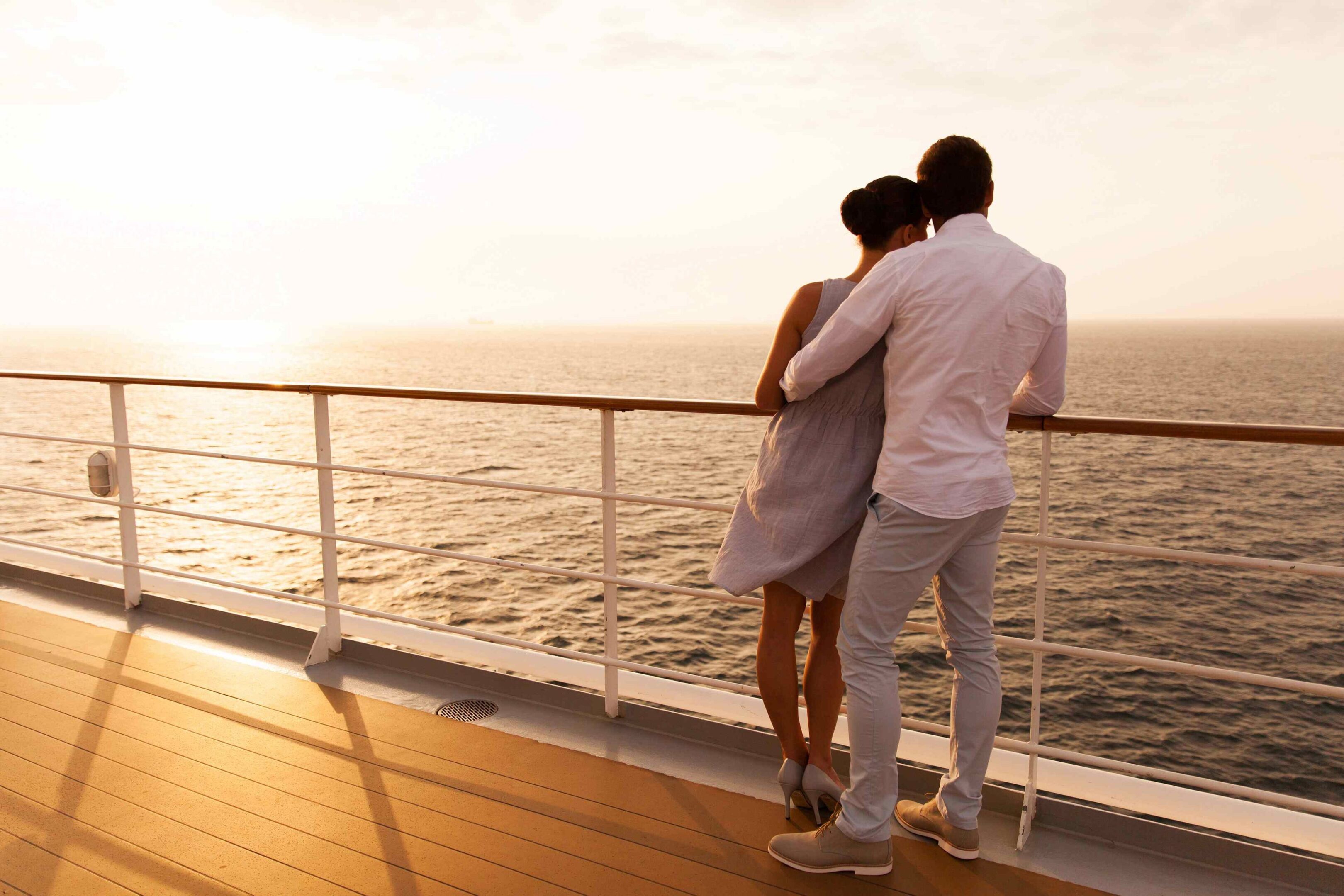 A couple stands on a ship's deck, gazing at the ocean during sunset.