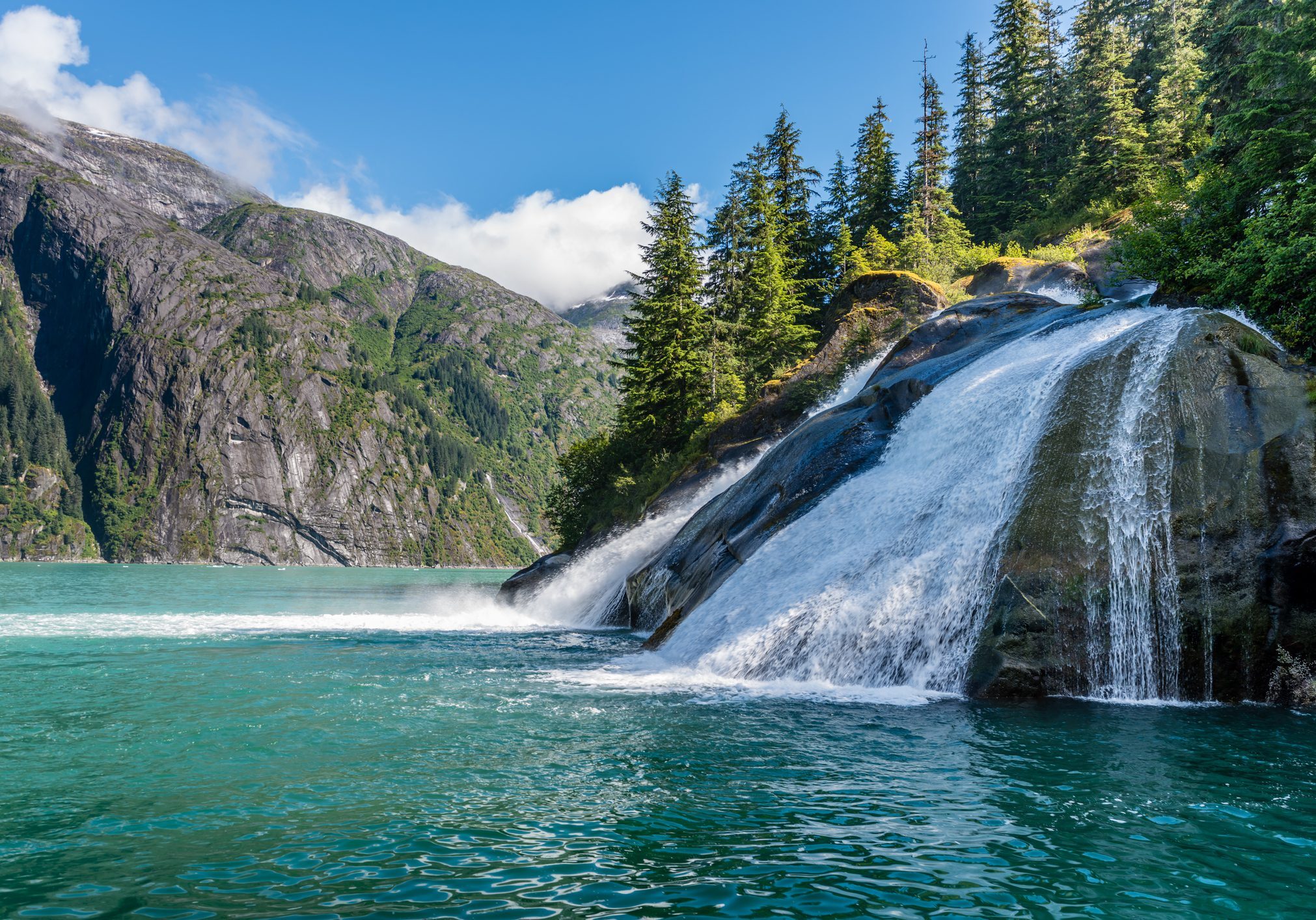 A serene waterfall flowing into a clear turquoise lake surrounded by pine trees and mountains.